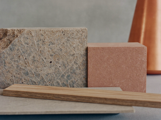 Nahaufnahme nebeneinandergelegter Materialproben: Naturstein, roher Putz, unglasierte Terrakottafliese und unbehandeltes Holz auf einer Betonfläche.
